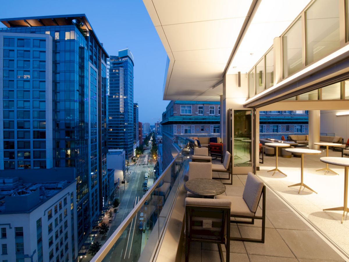 A modern urban balcony at dusk with outdoor seating overlooks a city street and tall buildings, illuminated by the surrounding lights.