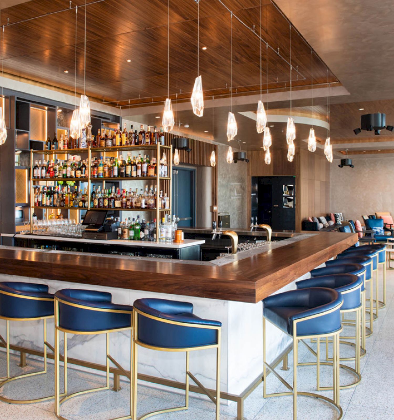 A modern bar with a large selection of liquors, surrounded by blue barstools with gold frames, and pendant lighting hanging from the ceiling.