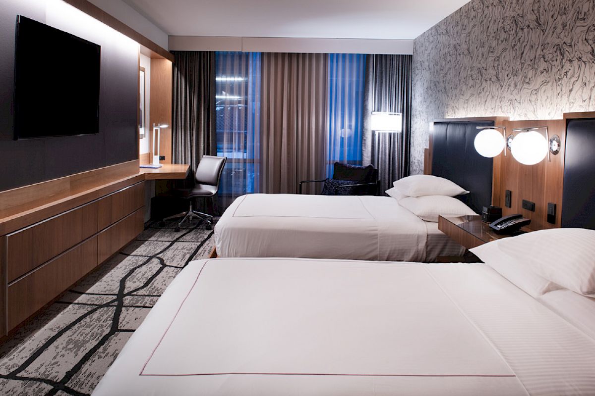 The image shows a modern hotel room with two neatly made beds, a wall-mounted TV, a desk and chair, and stylish lighting.