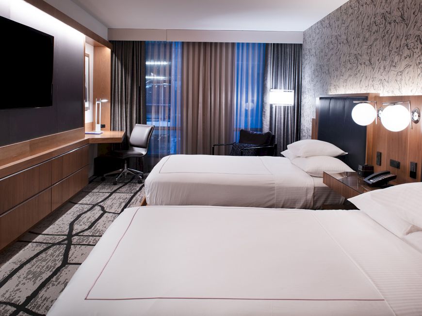 The image shows a modern hotel room with two neatly made beds, a wall-mounted TV, a desk and chair, and stylish lighting.