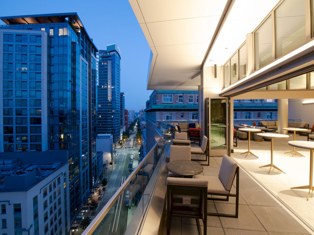 A modern cityscape at twilight, featuring a building with a spacious balcony that contains tables and chairs, overlooking a bustling street below.