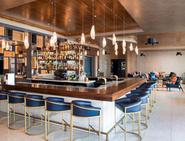 An upscale bar with blue chairs, hanging lights, and a fully stocked bar counter. The setting is modern with a cozy seating area in the background.