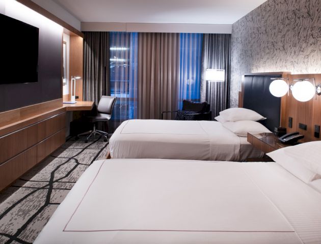 The image shows a modern hotel room with two neatly made beds, a flat-screen TV, a desk, chairs, and a large window with curtains.