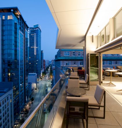 A modern balcony with furniture overlooks a cityscape of tall buildings and a street below, illuminated at dusk.