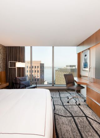 A modern hotel room with a large bed, flat-screen TV, desk, chair, and window offering a city and water view.