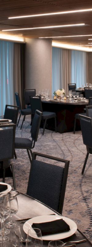 A modern banquet hall with round tables set for an event, featuring black chairs, floral centerpieces, and layered place settings under dim lighting.