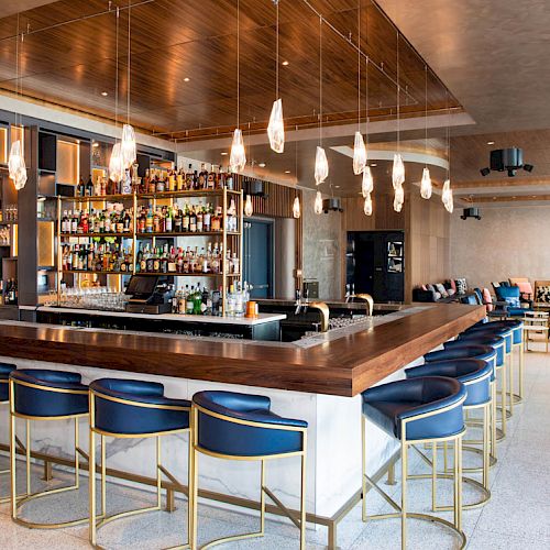 A modern bar with a wooden countertop, blue cushioned bar stools, hanging lights, and a well-stocked back bar, set in a spacious lounge area.