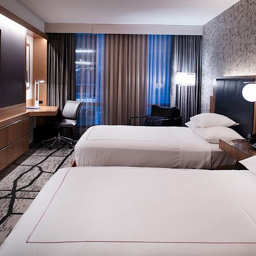 The image shows a modern hotel room with two neatly made beds, a wall-mounted TV, a desk and chair, and stylish decor and lighting.