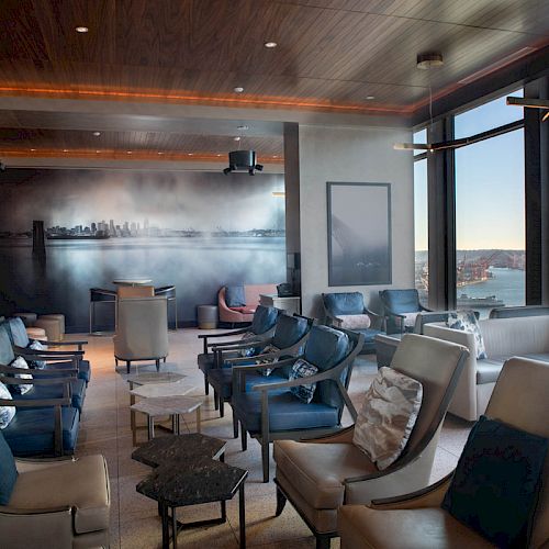 The image shows a modern lounge area with multiple chairs and tables, large windows with an ocean view, and a wall mural depicting a city skyline.