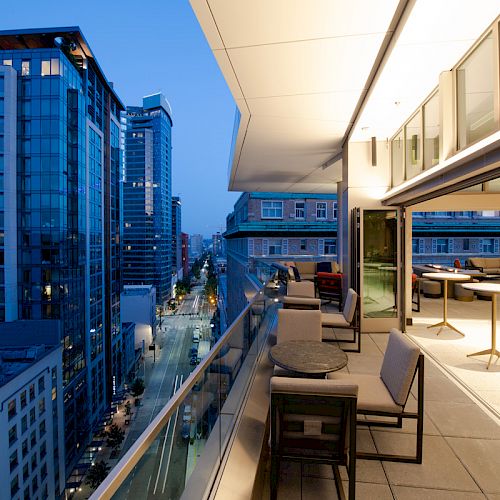 A modern balcony at dusk overlooking a cityscape with high-rise buildings, seating areas, and tables, offering a contemporary urban view.