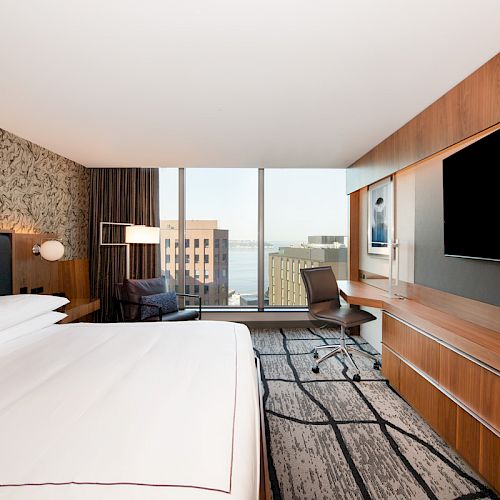 A modern hotel room with a large bed, a desk with a chair, a TV, and a window view of city buildings.