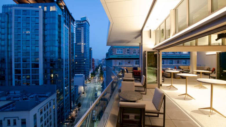 The image shows a modern balcony at dusk with chairs and tables, overlooking a city street surrounded by tall buildings and illuminated windows.