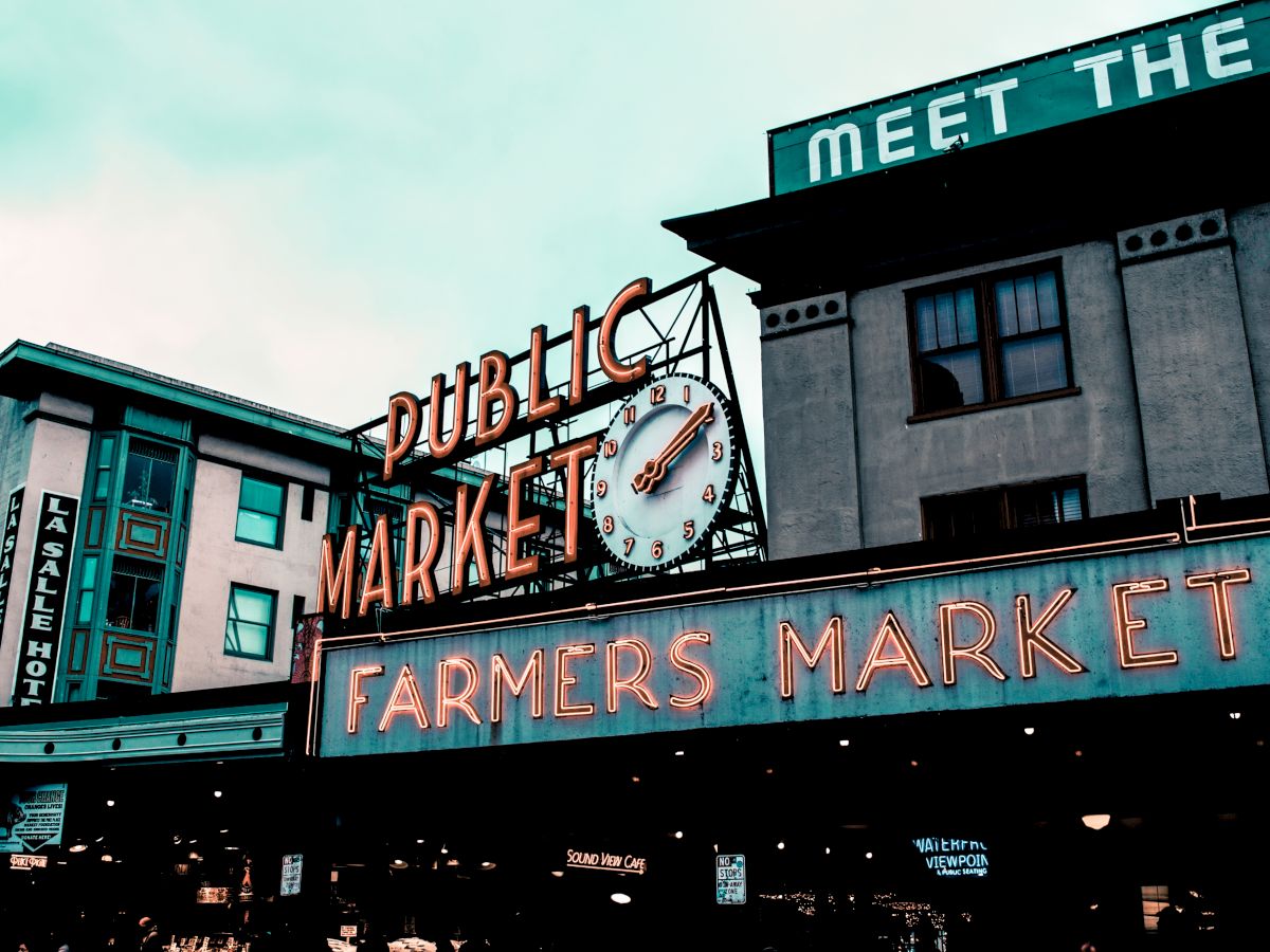 An image of a famous public market with neon signs reading "PUBLIC MARKET" and "FARMERS MARKET," featuring a clock, likely in Seattle.