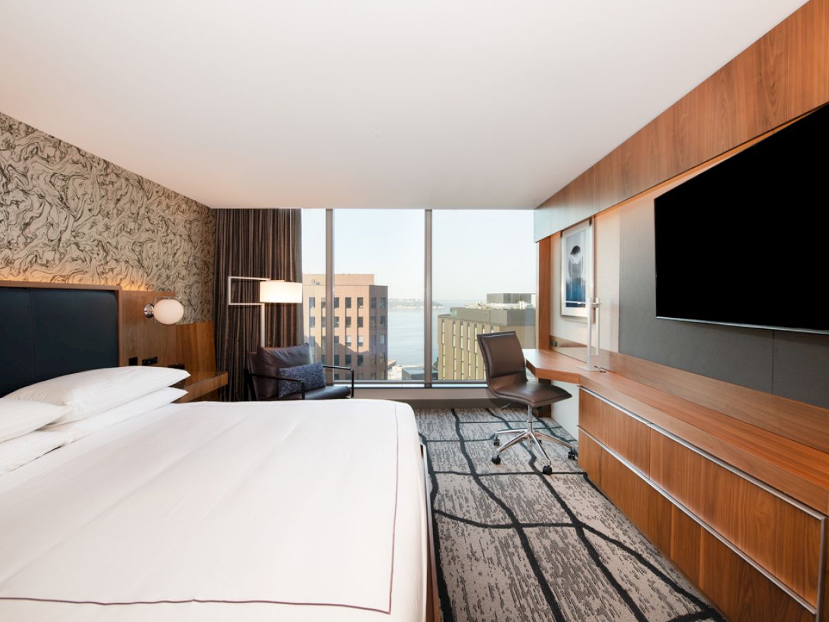 The image shows a modern hotel room featuring a large bed, a wall-mounted TV, a desk with a chair, and a window with a city view and some buildings visible.
