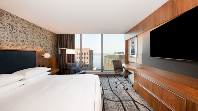 The image shows a modern hotel room featuring a large bed, a wall-mounted TV, a desk with a chair, and a window with a city view and some buildings visible.