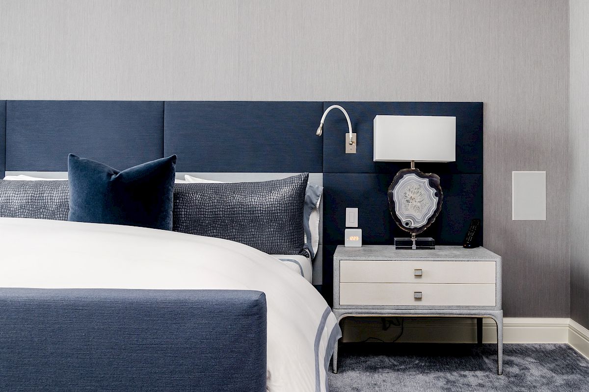 A modern bedroom features a bed with a blue headboard, white bedding, and gray cushions, alongside a nightstand with a lamp and decor.