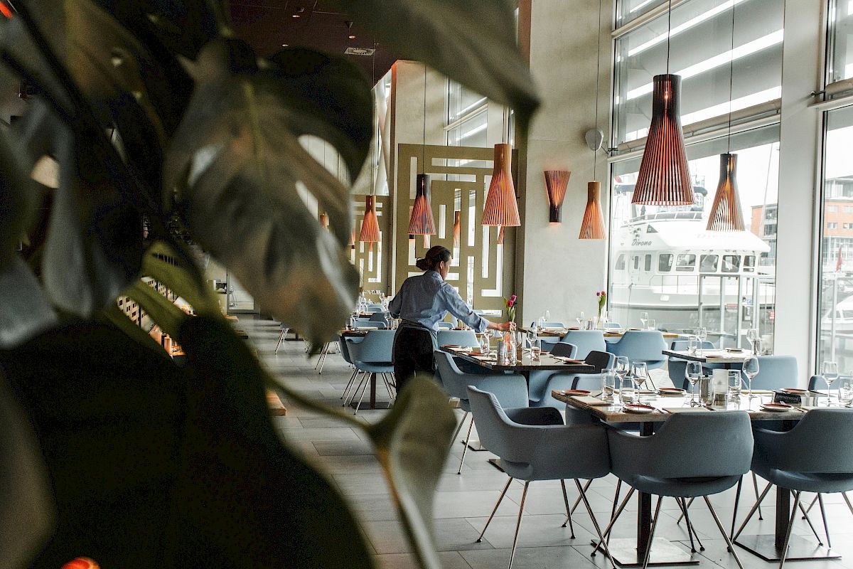 A person is setting up tables in a modern, stylish restaurant with large windows and unique lamps, partially obscured by leafy plants.