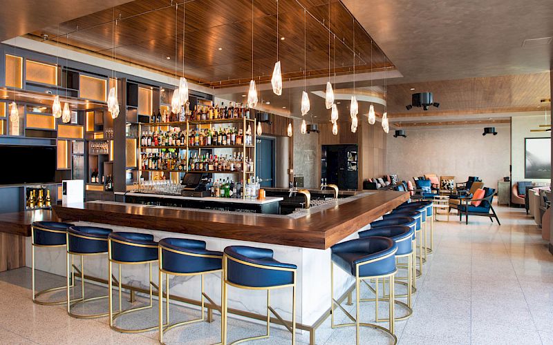 Modern bar with wooden accents, high shelves stocked with liquor, blue upholstered barstools, and hanging pendant lights.