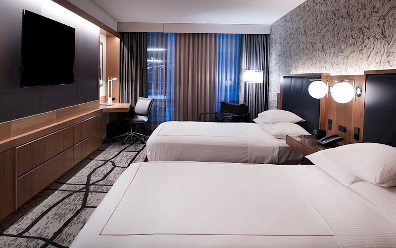 A modern hotel room with two beds, a flat-screen TV, a desk with a chair, and stylish lighting and decor.