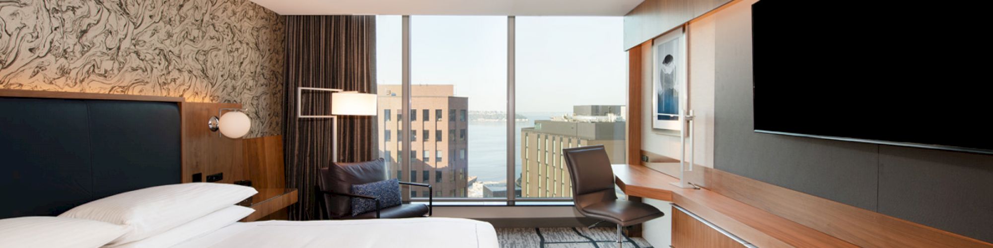 A modern hotel room featuring a large bed, a TV on a wooden console, a desk with a chair, and windows with a city view.